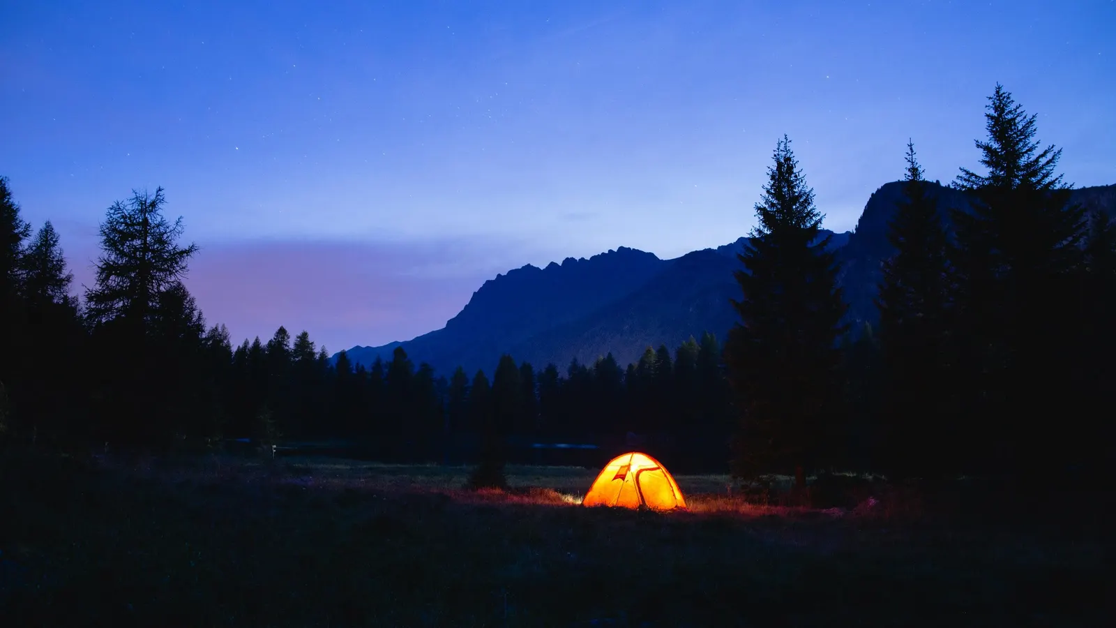 A tent lit from inside at dusk in a clearing, with pine trees and a mountain behind